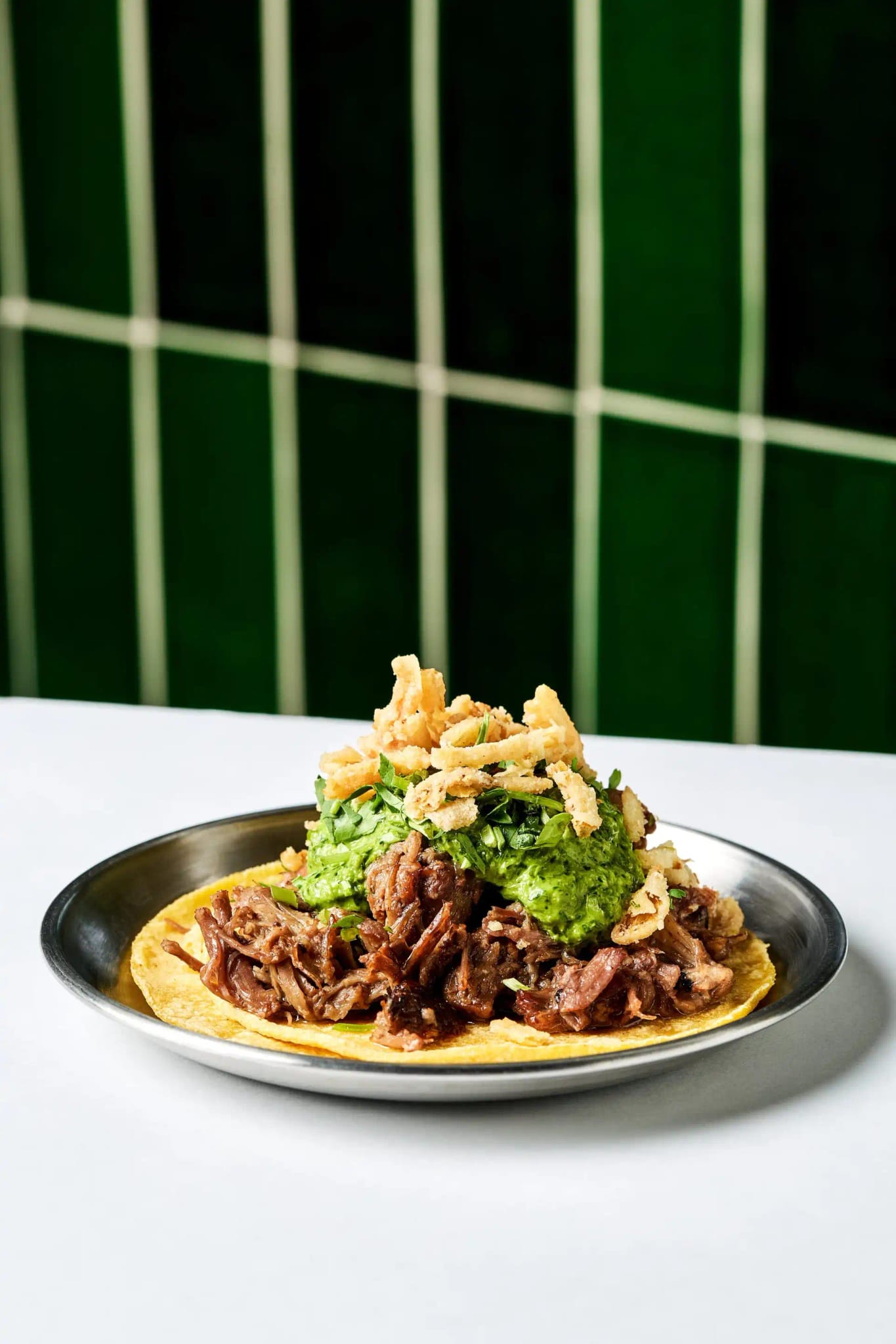 A crisp tostada piled with slow-cooked meat, guacamole and fried onions on a steel plate in front of Lenny's green tile wall