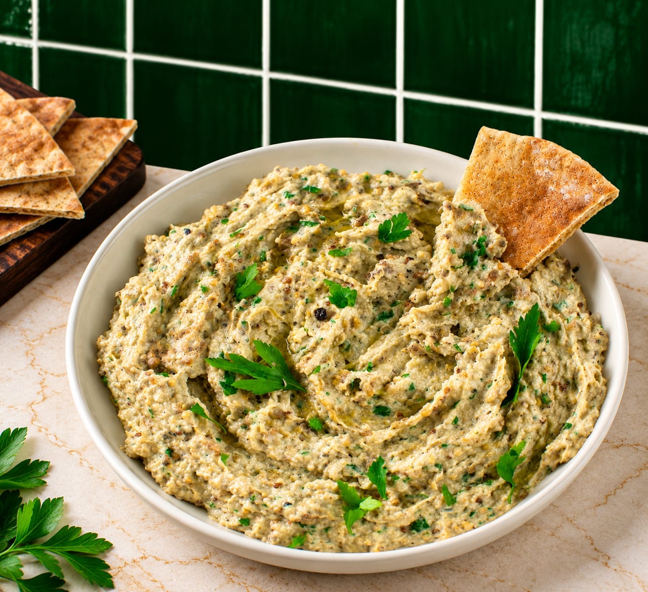 Babaganoush swirled in a white bowl with parsley and a pita chip, pita wedges on a board beside it