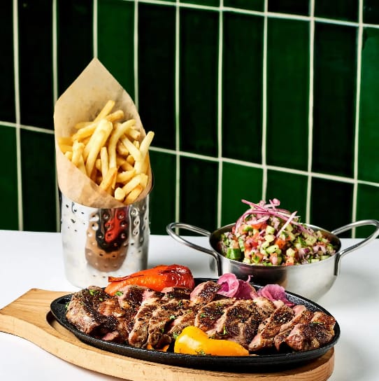 Sliced rib eye with grilled peppers and pickled red onions on a skillet, beside French fries and a pan of Israeli salad, against green tile