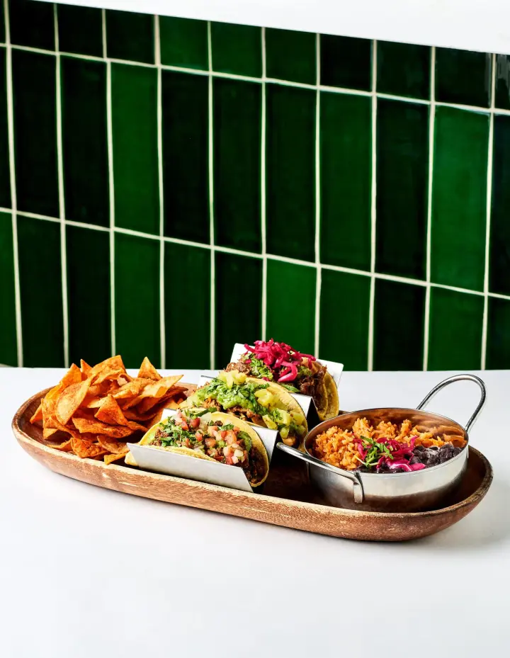 Three tacos in steel holders with chips, rice and black beans on a wooden board