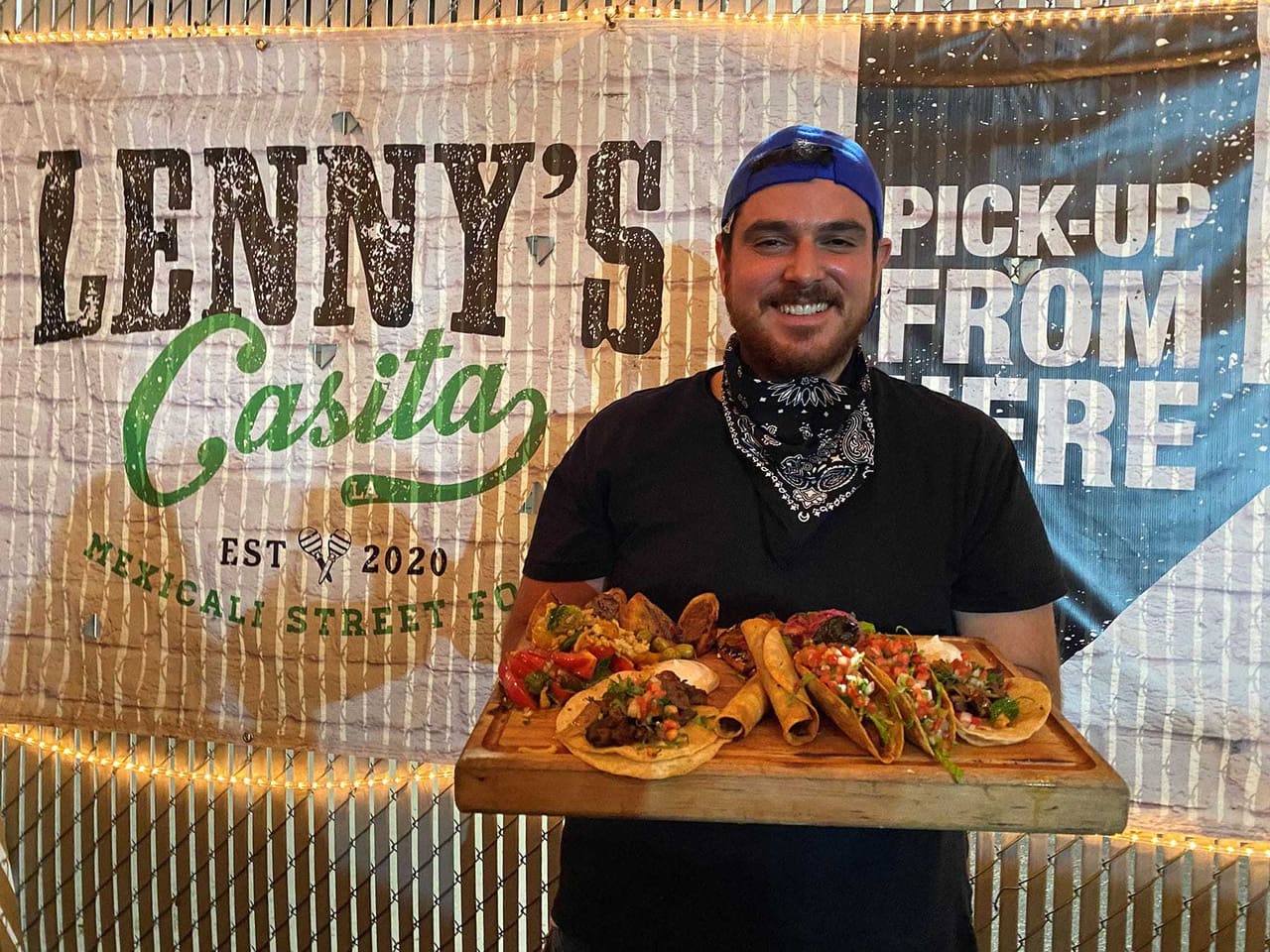 Chef Lenny smiling in a blue cap and bandana, holding a wooden board of tacos, flautas and sides in front of the Lenny's Casita banner