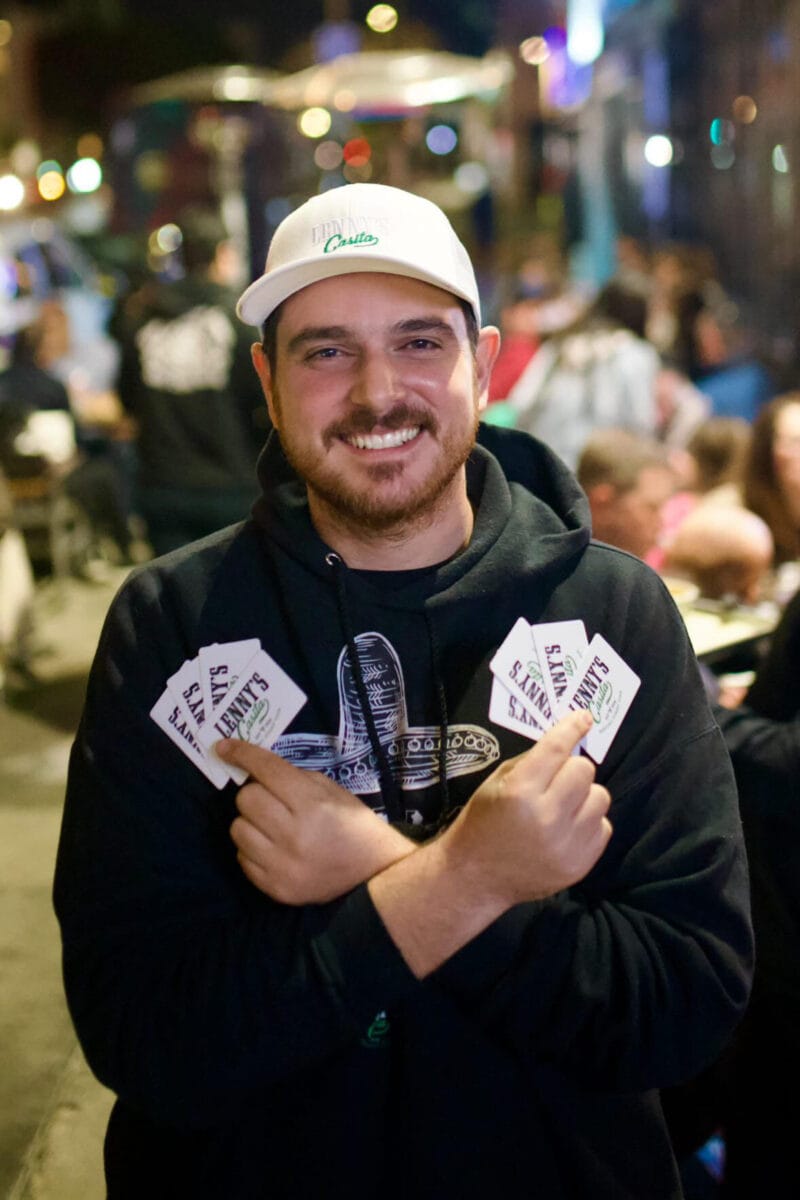 Chef Lenny in a white Casita cap and black hoodie, smiling with his arms crossed at a crowded night event