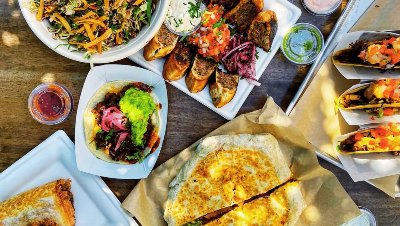 Overhead of takeout plates in the sun: a guacamole taco, grilled kebab rolls with salsa, a crisp salad, hard-shell tacos and a pressed burrito