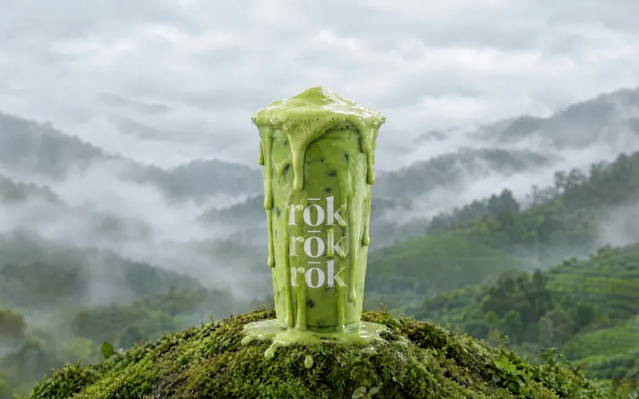 A matcha latte in a clear cup printed with rōk, overflowing onto moss above misty tea hills.