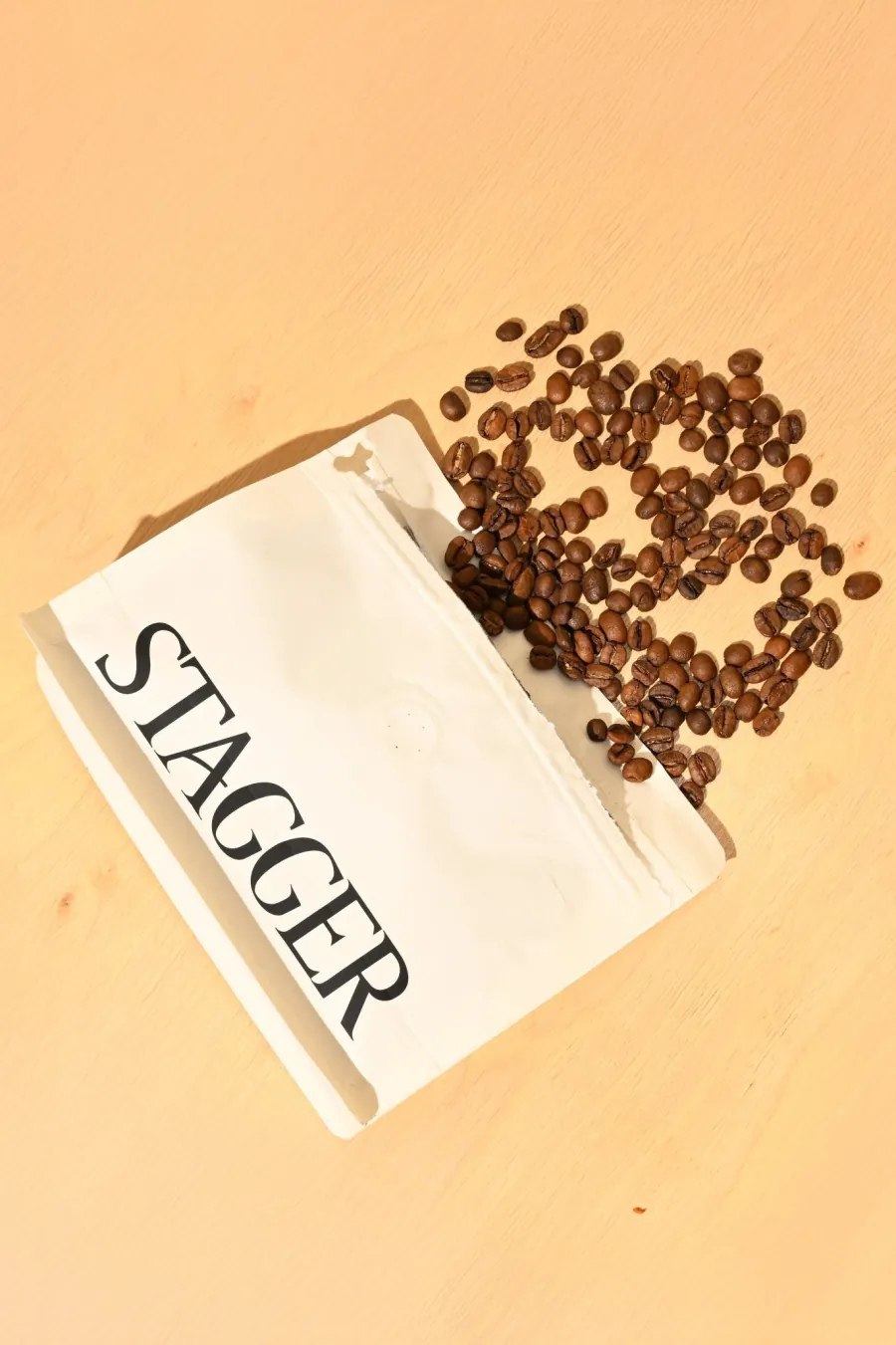 A white Stagger coffee bag spilling whole roasted beans across a warm surface