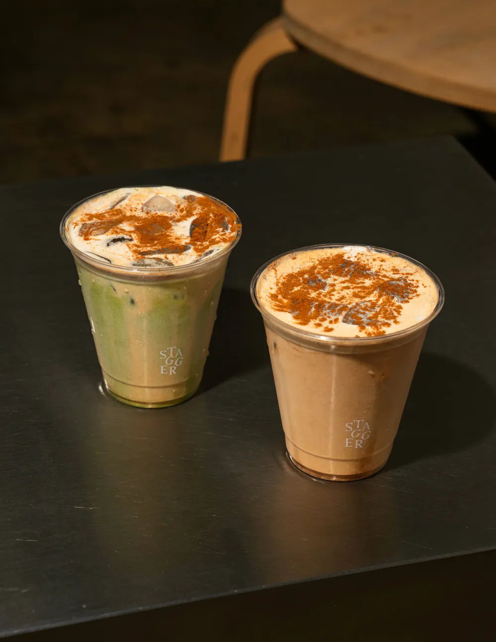 Two iced drinks dusted with cinnamon on a black metal table, a bentwood chair behind