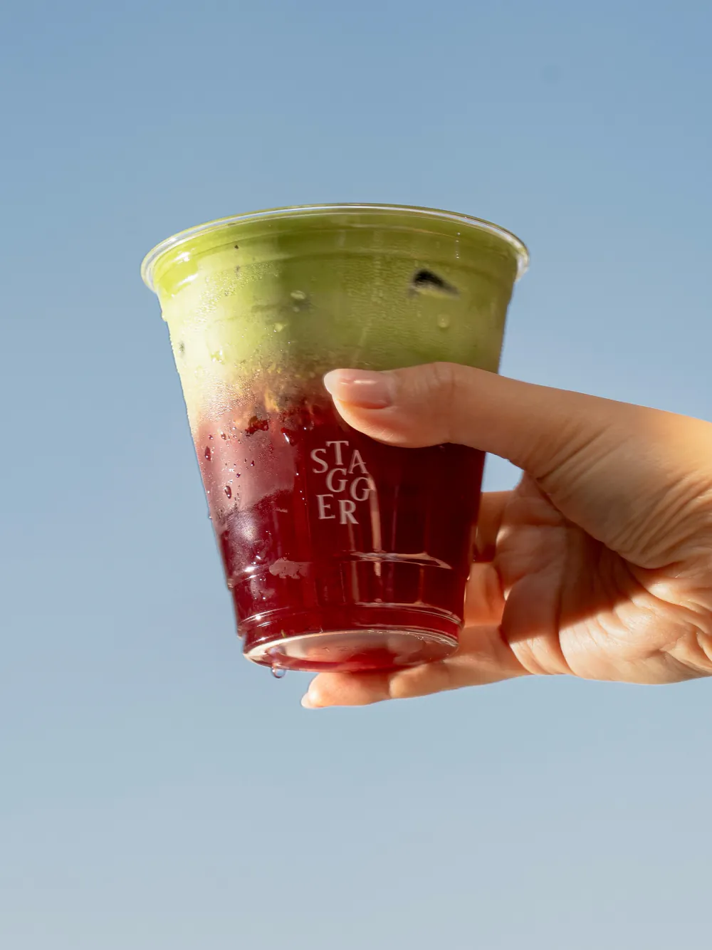 A layered green and crimson Stagger drink held up against a clear blue sky