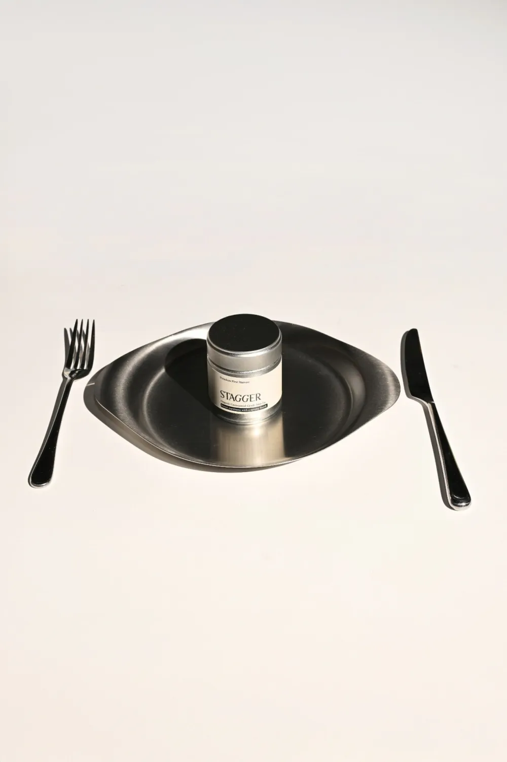 A Stagger matcha tin set on a steel plate between a fork and knife
