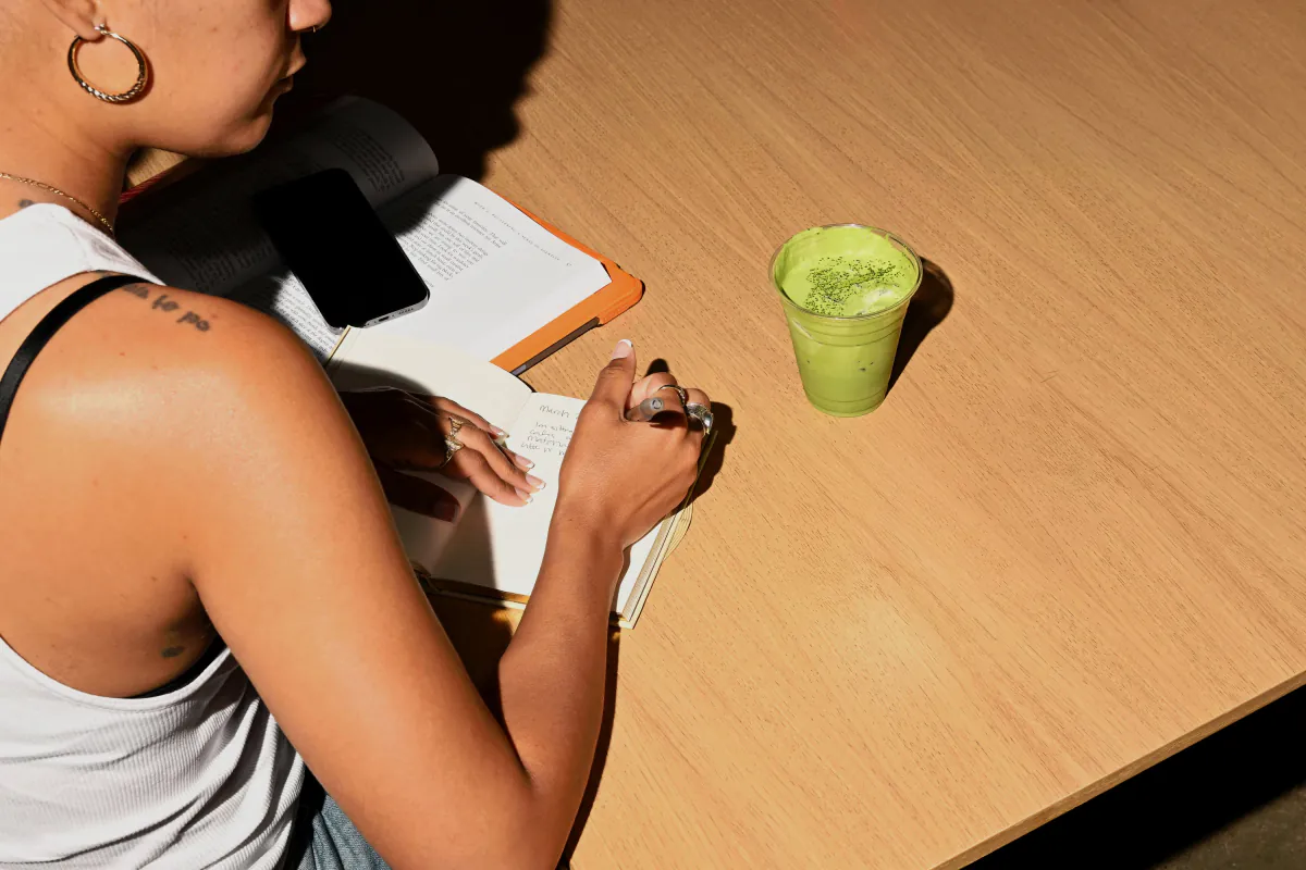 A guest writes in a notebook at a sunlit wooden table beside an iced matcha latte